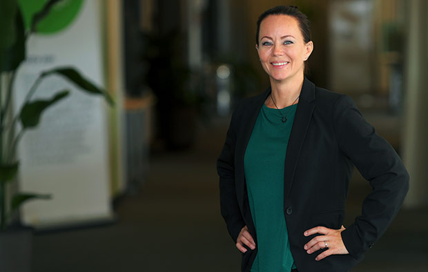 Maria Monthure står i ett kontorslandskap på Bolagsverket. Hon har svart kavaj och grön tröja. I bakgrunden skymtar en blomma och kontorsmöbler. 