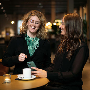 Två personer står vid ett bord och tittar i mobilen och skrattar. På bordet står det en kopp kaffe och ett värmeljus.