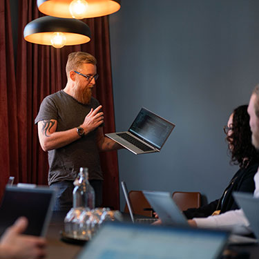 En person med en laptop i ena handen pratar med en grupp personer som sitter runt ett bord.