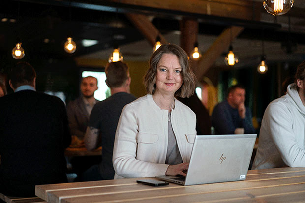 En kvinna med axellångt, mörkblont hår sitter vid en bärbar dator av märket HP på ett träbord i en modern, mysigt upplyst miljö. I bakgrunden syns flera personer i samtal vid andra bord, med dekorativa glödlampor som hänger från taket.