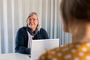 En kvinna med glasögon sitter vid en bärbar dator och tittar på en annan person i förgrunden. Kvinnan bär en mörk tröja och en grå scarf. De samtalar i en ljus kontorsmiljö med ljusa gardiner i bakgrunden.