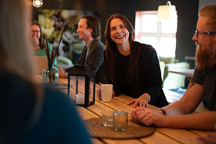 A group of colleagues sitting around a wooden table in a cosy, warmly lit room, engaged in cheerful conversation. A woman in the center is smiling brightly, holding a mug, while others around her listen and laugh.