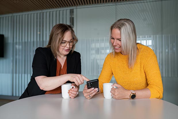 Två kvinnor sitter vid ett runt bord inomhus. Den ena kvinnan, med glasögon och mörk kavaj, pekar på en mobiltelefon som den andra kvinnan, klädd i en gul stickad tröja, håller i handen. Båda ler och har varsin kaffekopp framför sig. I bakgrunden syns en glasvägg med lodräta ränder.