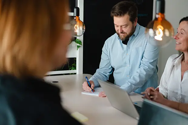 Tre personer samtalar vid ett bord. En man antecknar medan en kvinna sitter framför en bärbar dator.