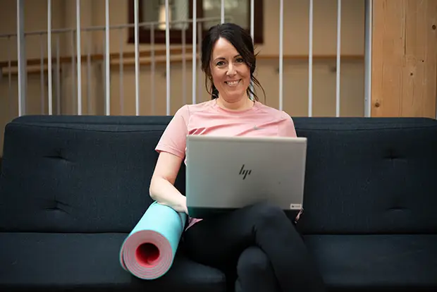 Kvinna sitter i en soffa med en bärbar dator i knät och en yogamatta bredvid sig.