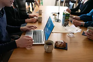 Flera personer sitter runt ett bord med kaffekoppar och fikabröd. En person arbetar på en bärbar dator medan de andra samtalar.
