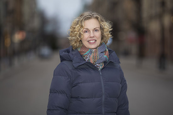 En person står i utomhusmiljö, klädd i en mönstrad halsduk och en mörkblå täckjacka.