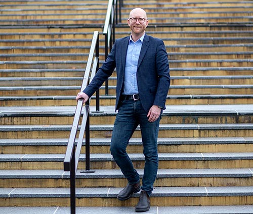 En person står och håller i ett räcke i en trappa utomhus. Personen är klädd i en blå skjorta, mörkblå kavaj, mörkblå jeans och bär glasögon.