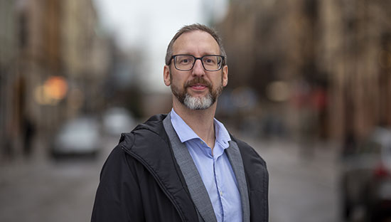 En person står i utomhusmiljö klädd i en ljusblå skjorta, mörk jacka och bär glasögon.