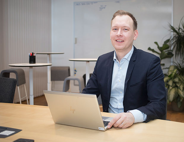David Magård sitter vid ett bord på Bolagsverket. Han har blå kavaj och ljusblå skjorta och arbetar med dator. I bakgrunden syns en vit tavla och en grön växt.