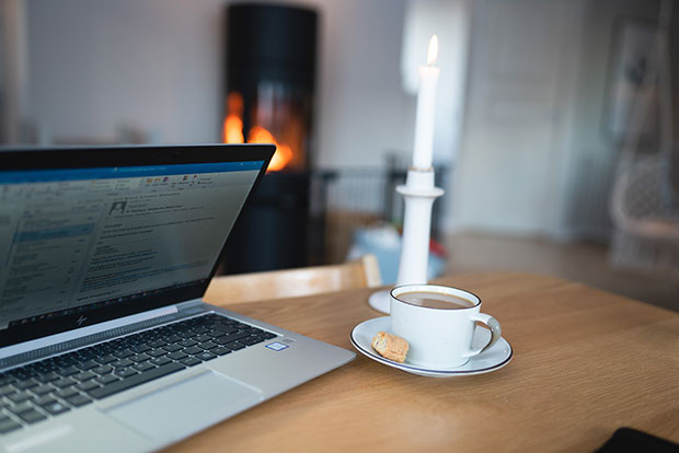 En bärbar dator med ett öppet e-postprogram står på ett träbord. Bredvid datorn finns en kopp kaffe på ett fat med ett litet kex samt en tänd stearinljus. I bakgrunden syns en braskamin med synliga lågor, vilket skapar en varm och hemtrevlig atmosfär.