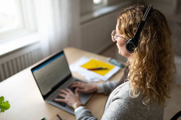 Kvinna sitter vid skrivbord framför fönster med laptop framför sig.