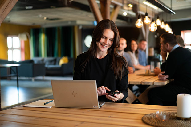 En glad tjej sitter vid ett långt träbord och arbetar på sin laptop samtidigt som hon använder sin mobiltelefon. I bakgrunden syns fler personer som samtalar i en modern kontorsmiljö med mysig belysning och trädetaljer.