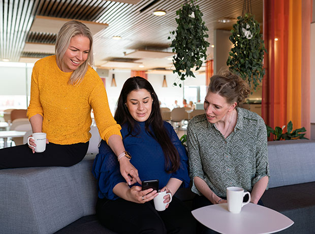 Tre kvinnor sitter i en lounge-miljö med kaffekoppar i händerna. Kvinnan till vänster, klädd i en gul tröja, lutar sig fram och pekar på en mobiltelefon som kvinnan i mitten, klädd i blått, håller. Kvinnan till höger, i en grön mönstrad blus, tittar också på skärmen. I bakgrunden syns gröna växter som hänger från taket och ett ljust, modernt kontorslandskap