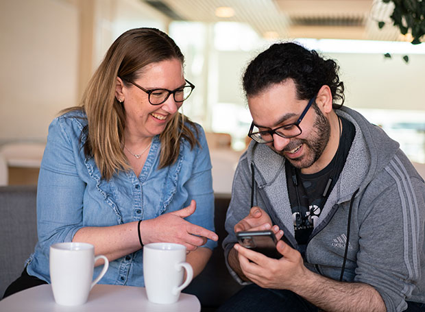 En kvinna och en man sitter tillsammans vid ett bord med två kaffekoppar. Kvinnan, som bär glasögon och en blå blus, ler och pekar på en mobiltelefon som mannen, klädd i en grå huvtröja med glasögon, håller. Båda ser glada ut i en ljus och modern inomhusmiljö.
