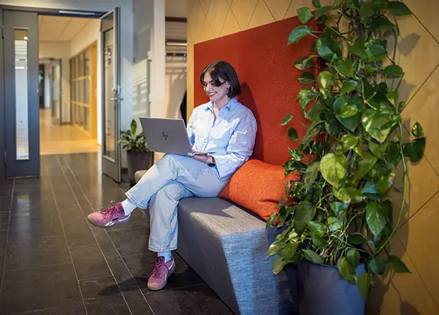 Ann Johansson sitter på en bänk i en kontorsmiljö och arbetar på en bärbar dator. Hon lutar sig mot en röd vägg, har ljusa kläder och rosa skor. Bredvid finns gröna växter och en korridor sträcker sig in i bakgrunden.
