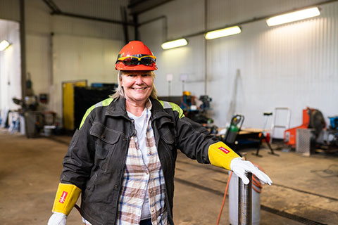 Kvinna i en industriell verkstadsmiljö bär skyddsutrustning, inklusive röd hjälm med skyddsglasögon, gula skyddshandskar och en svart arbetsjacka över en rutig skjorta. Hon ler mot kameran och vilar ena handen på ett metallrör. I bakgrunden syns maskiner och verktyg i en rymlig verkstad med vita väggar.