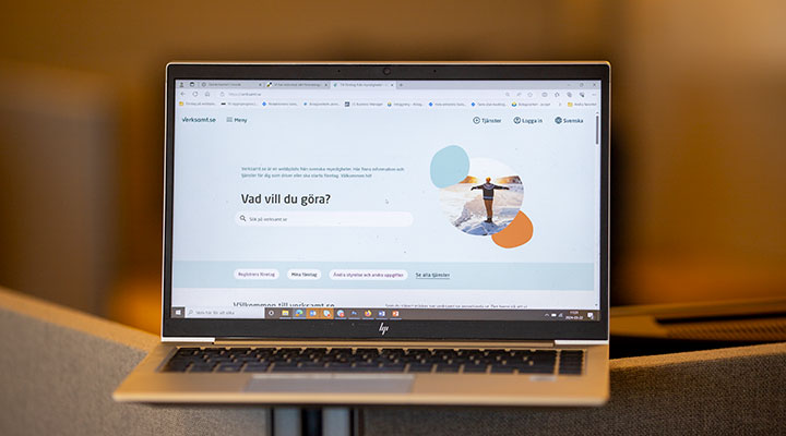 En bärbar dator med en öppen webbsida som visar startsidan för verksamt.se. På skärmen syns en sökruta med texten 'Vad vill du göra?' och en bild på en person med utsträckta armar vid havet. Datorn står på ett grått bord i en inomhusmiljö med suddig bakgrund.