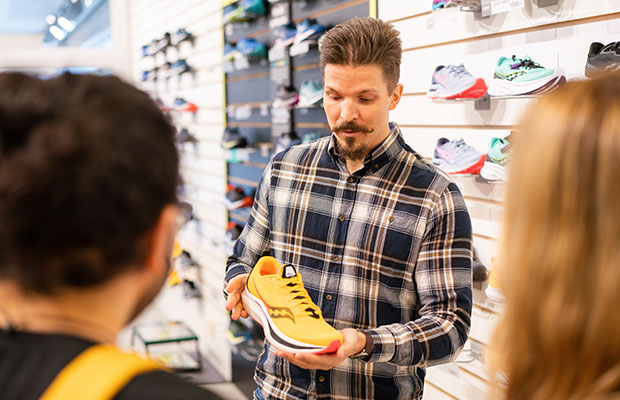 En butikspersonal i rutig skjorta visar upp en gul träningssko för kunder i en skobutik. Skorna är uppställda på hyllor i bakgrunden.