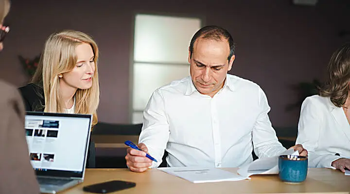 Several people are sitting in a meeting with papers and laptops on the table in front of them.