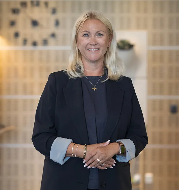 Porträtt av Sarah Eklund som står inomhus och ler mot kameran. I bakgrunden syns en vägg med träpanel och en väggklocka.