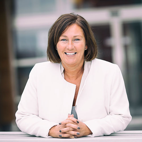 Nina Brede. Med axellångt brunt hår ler mot kameran. Hon bär en vit kavaj över en grå topp och har händerna vilande framför sig. Bakgrunden är suddig med inslag av fönster och byggnadsdetaljer i ljusa toner.