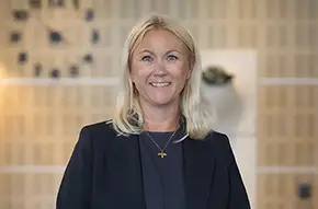 Porträttbild av Sarah Eklund, klädd i mörk kavaj och mörk topp, stående inomhus framför en ljus bakgrund med suddiga detaljer.