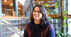 En glad person med mörkt långt hår och glasögon sitter i ett stråk i Bolagsverkets byggnad.