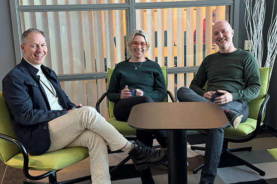 It-produktions sektionschefer sitter avslappnat i gröna fåtöljer runt ett bord med en kopp kaffe i händerna. 