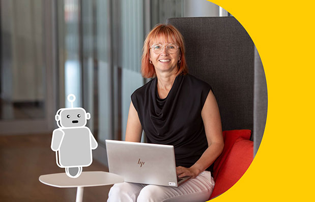 En kvinna sitter vid ett litet bord med en bärbar dator framför sig. På bordet står en tecknad robot.