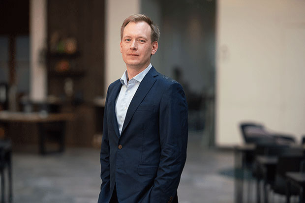 En
man i en mörkblå kostym och ljusblå skjorta står i en modern kontorsmiljö. Han
har kort, ljust hår och ett neutralt ansiktsuttryck. Bakgrunden är suddig och
visar bord och stolar i en öppen arbetsyta.