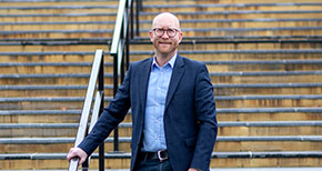 Leende man i kostym står vid ett trappräcke utomhus.