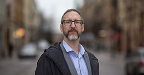 Man med glasögon står utomhus på en gata, tittar in i kameran.