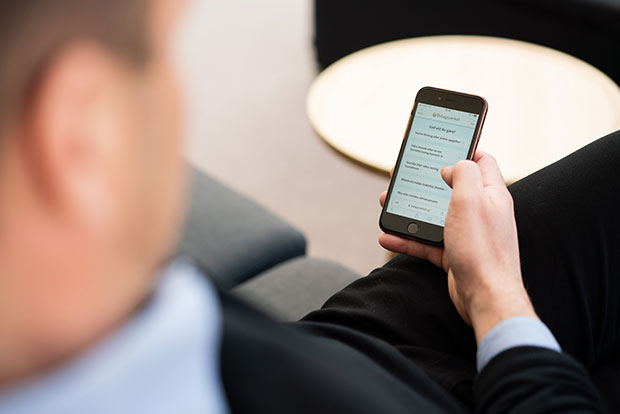 En person i kostym sitter i en soffa och håller i en mobiltelefon. På mobilskärmen syns en webbsida med text