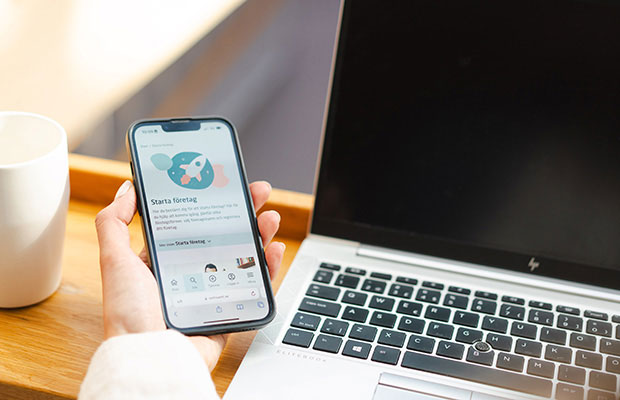 En hand håller i en mobiltelefon som visar sida från verksamt.se på skärmen. I bakgrunden syns en kaffekopp och en laptop.