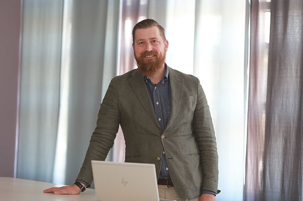 Martin Roos står vid ett bord och lutar ena handen mot det. Framför honom står en öppen bärbar dator. Han bär en kavaj över en skjorta och ler mot kameran. I bakgrunden syns ljusa gardiner i olika färgtoner.