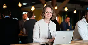 A woman works on a laptop in a setting with other people in the background.