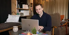 A man sits on a sofa working on a laptop in a home environment.