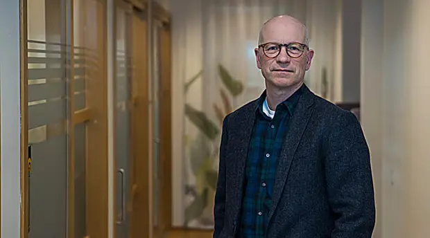Ola Pettersson med glasögon, står i en kontorsmiljö. Han bär en mörk kavaj över en mörkgrönrutig skjorta och tittar in i kameran. I bakgrunden syns glasväggar, dörrar och växter i mjukt fokus.