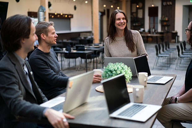 Flera personer sitter runt ett bord med sina laptops och pratar.