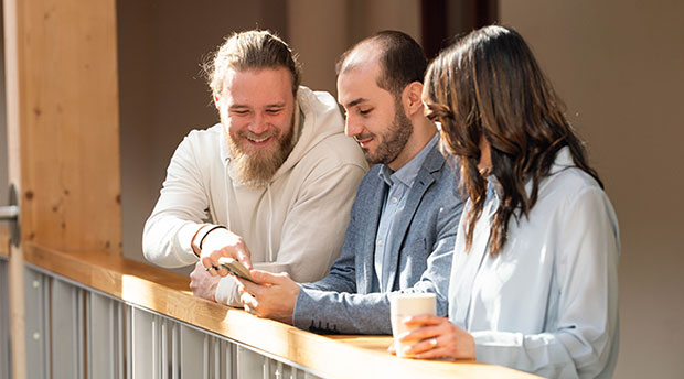 Tre personer står vid ett räcke inomhus. De tittar tillsammans på en mobiltelefon och ler. En person håller en kaffemugg.